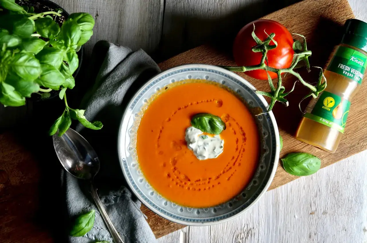 En sk&aring;l med kr&auml;mig tomatsoppa, gjord p&aring; krossade tomater, toppad med en klick gr&auml;ddfil och basilika. En f&auml;rsk tomat och en flaska Knorr Fresh Vegetable Vegan Fond ligger bredvid.