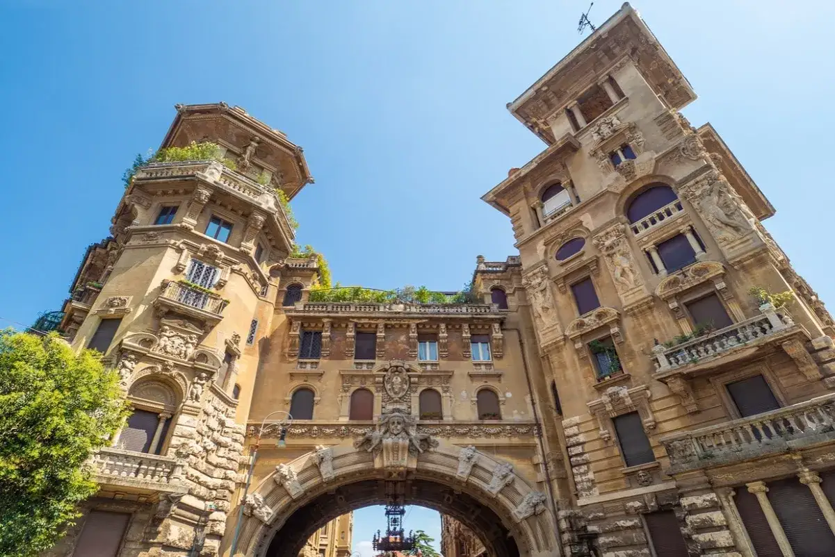 Edifici fiabeschi con torrette e decorazioni elaborate nel quartiere Copped&egrave;, Roma. Un arco monumentale collega le strutture sotto un cielo azzurro.