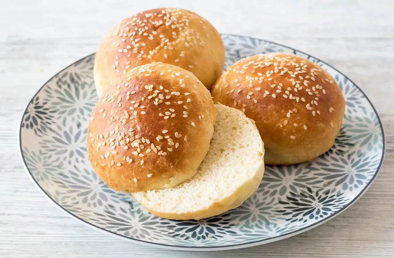 Tre soffici burger roll cosparsi di semi di sesamo, uno tagliato a metà, su un piatto decorato.