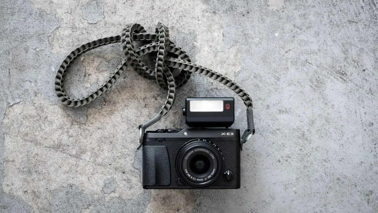 Czarny aparat Fujifilm X-E3 z obiektywem i lampą błyskową, przewieszony przez pleciony pasek, leży na szarej, popękanej powierzchni.