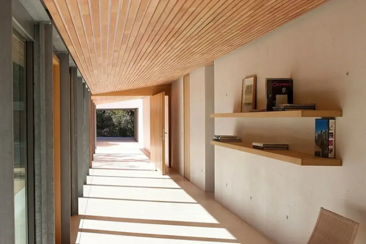 Corridoio moderno con soffitto a listelli in legno, pareti in cemento grezzo e mensole con libri. La luce solare crea un gioco di ombre sul pavimento.
