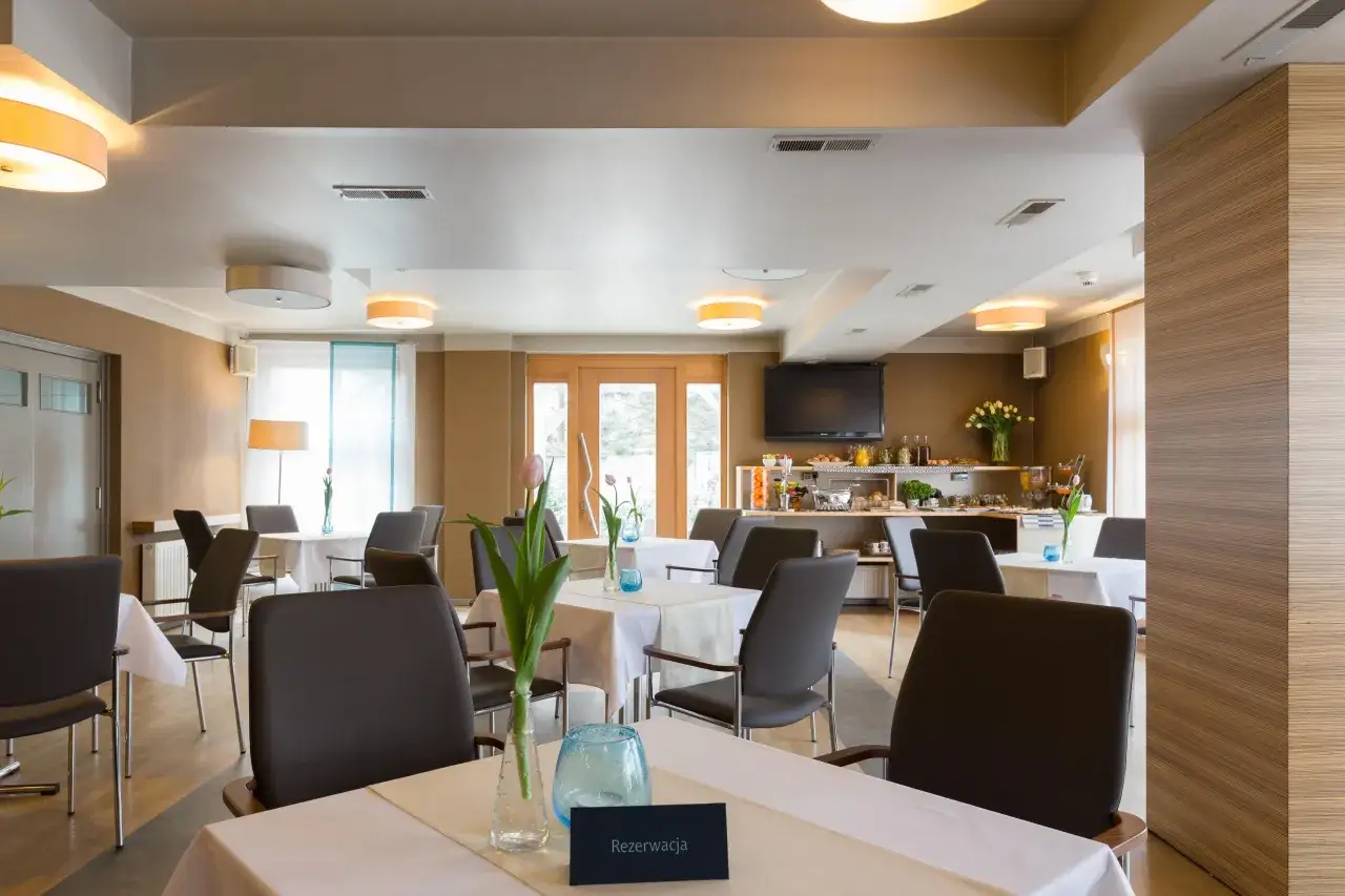 Elegancka sala restauracyjna w hotelu Perła Kraków. Stoły nakryte białymi obrusami, krzesła z ciemnej skóry, w wazonach tulipany.