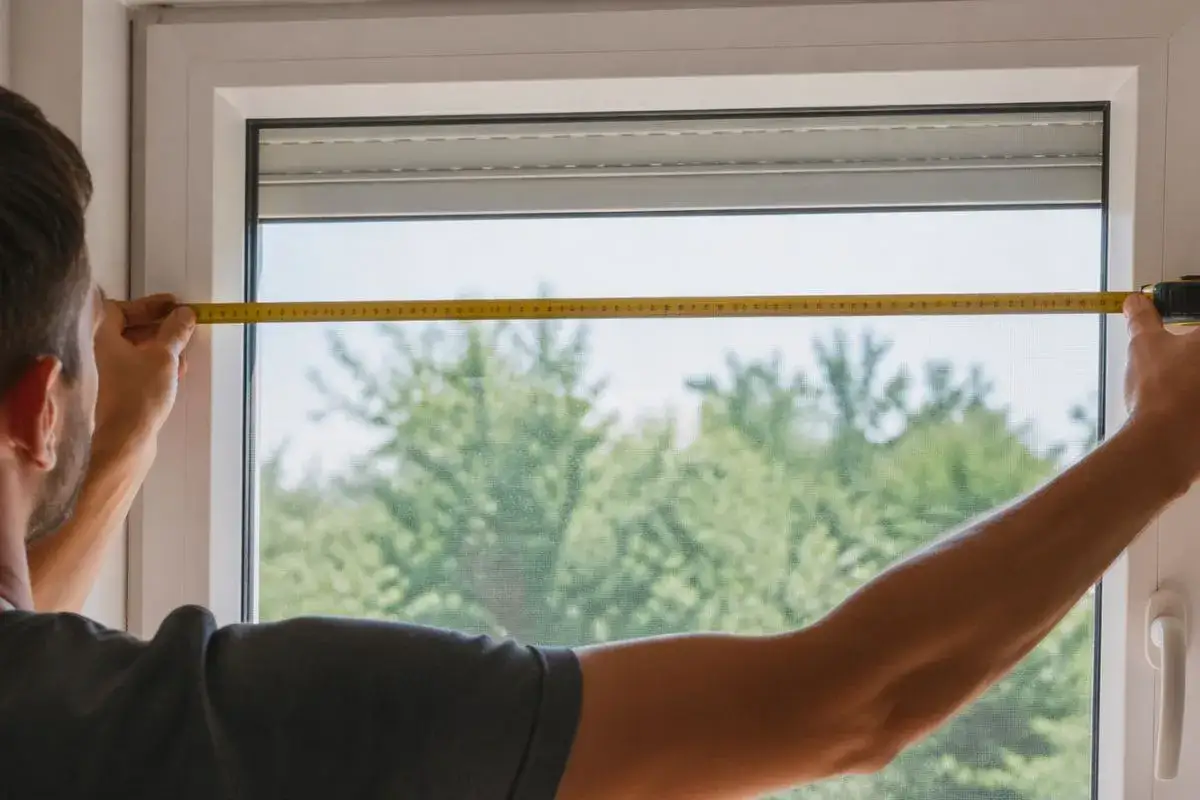 Un homme mesure une fen&ecirc;tre avec un m&egrave;tre ruban. Il est essentiel de savoir comment mesurer une fen&ecirc;tre pour installer de nouveaux rideaux ou stores.