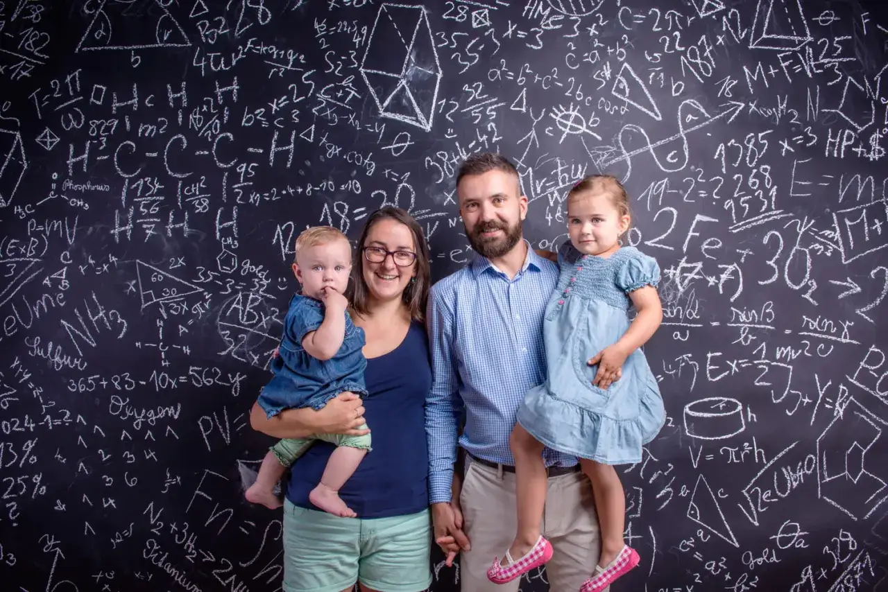 Famille souriante devant un tableau noir rempli d'équations. Une mère tient un bébé, un père tient une fillette. Une belle illustration de la vie avec un HPI, où la curiosité et l'intelligence se côtoient.