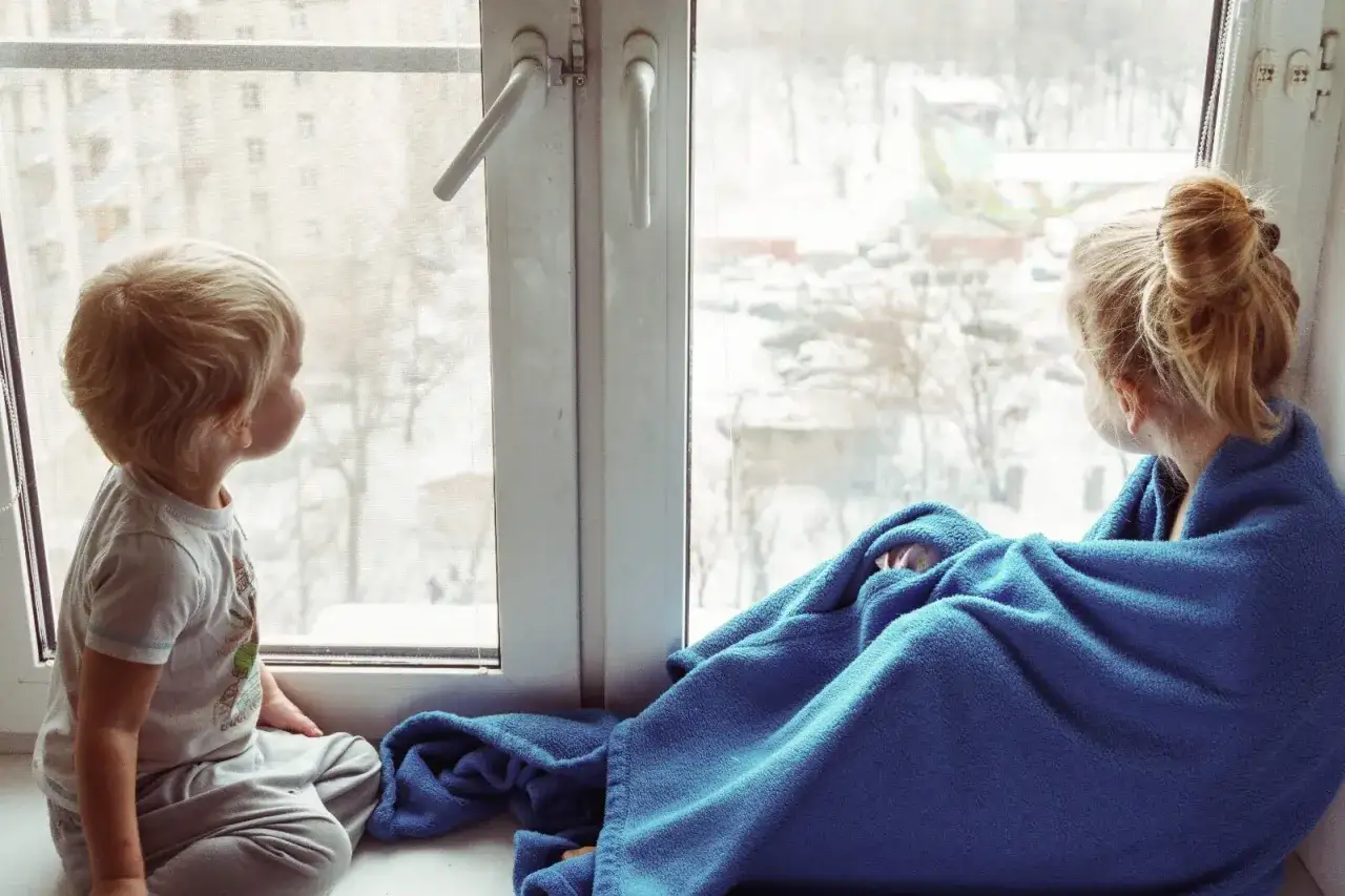 Dos niños, uno de 3-4 años, observan la nieve desde la ventana. Uno está envuelto en una manta azul.