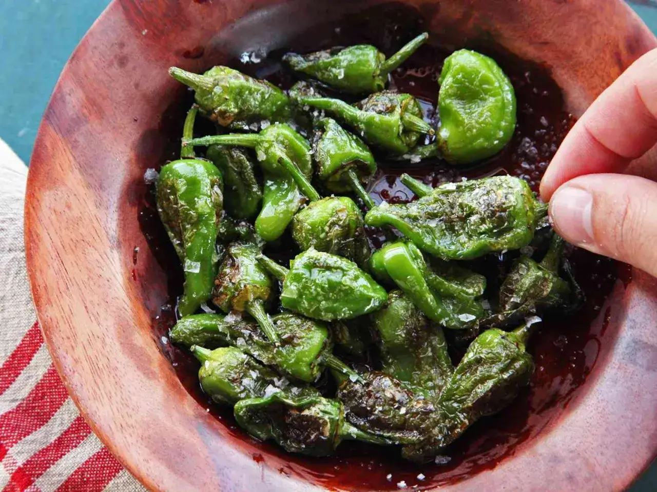 En sk&aring;l med friterade pimientos de padron, str&ouml;dda med flingsalt. En hand plockar en peppar.