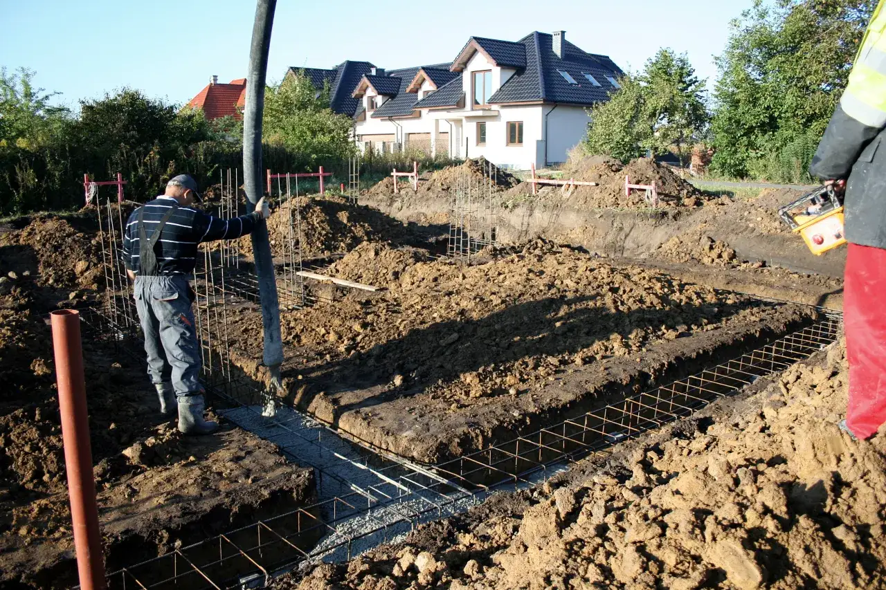 Pracownik wylewa beton do wykopu pod ławy fundamentowe. W tle widać domy w budowie.