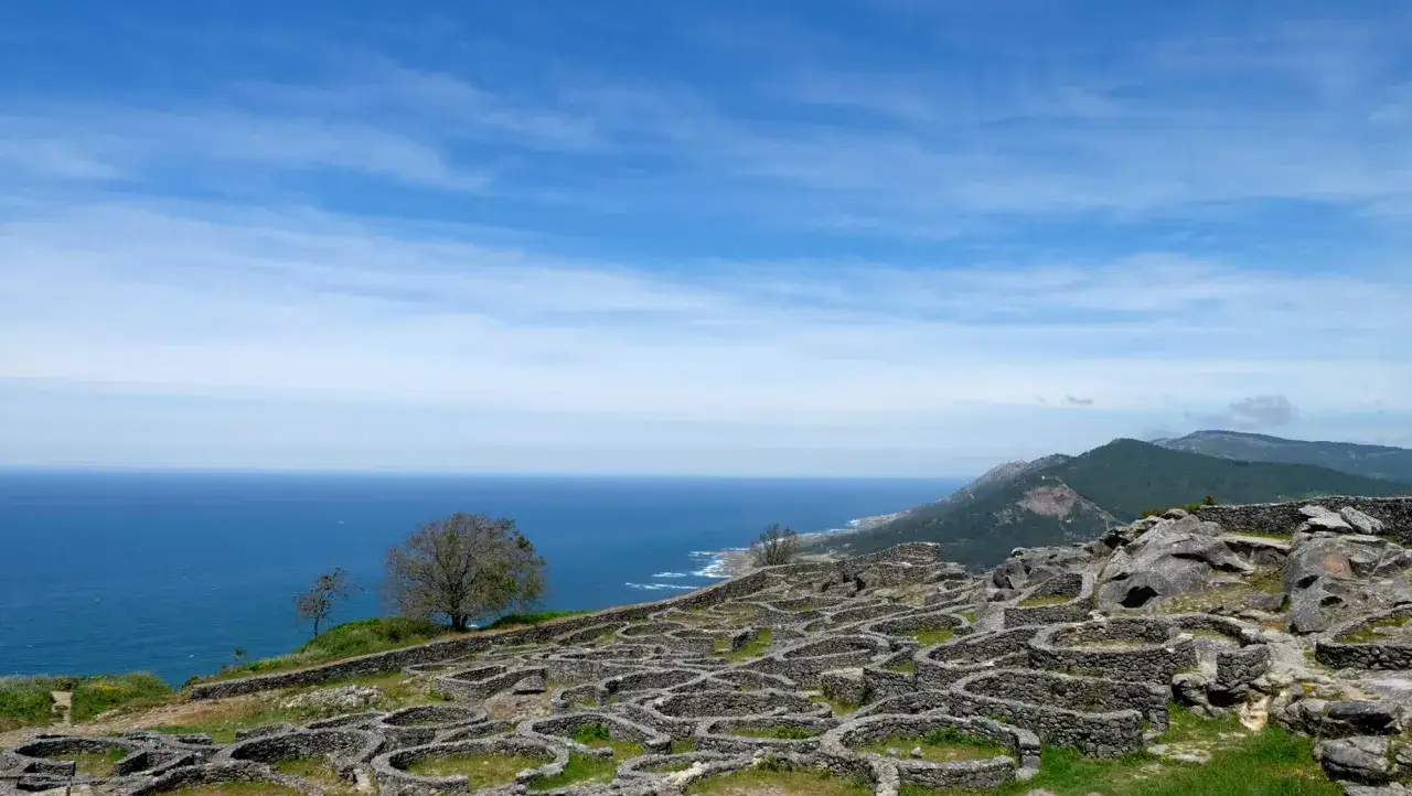 Ruinas circulares de piedra en el mirador Santa Tecla, Galicia, con el mar azul y montañas verdes al fondo.