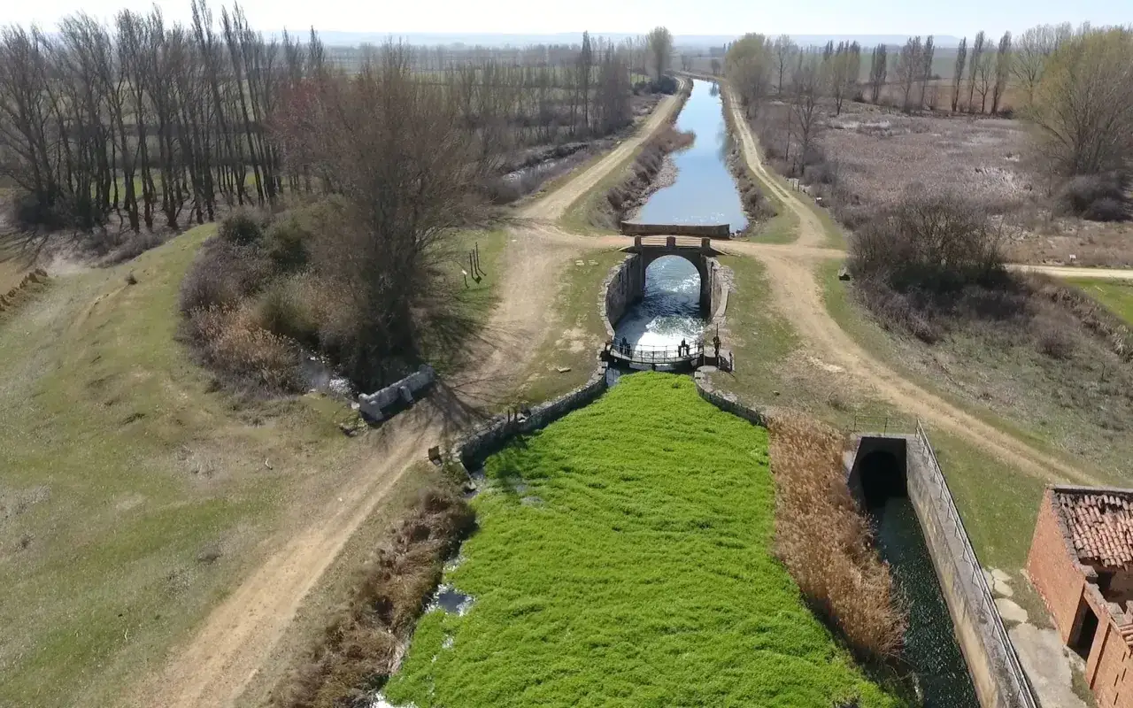 Canal de agua con vegetación verde exuberante, un puente y caminos de tierra cerca de Boadilla del Camino.