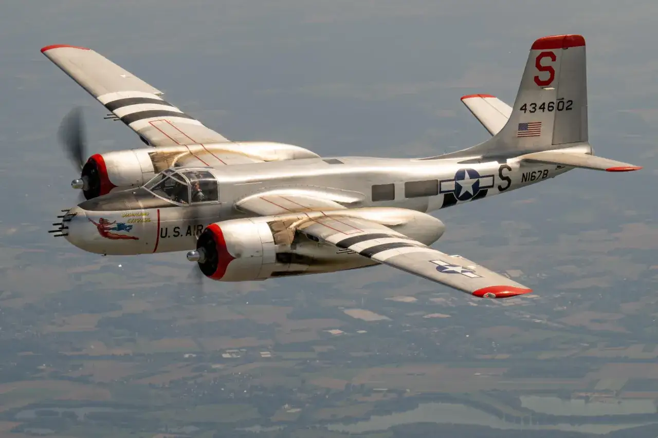 Silberne A-26 Invader mit roten Motorkapseln und schwarzen Streifen auf den Flügeln fliegt über eine Landschaft.