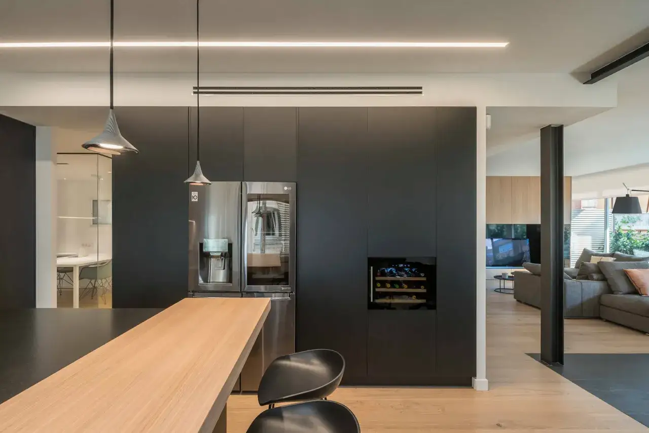 Cocina moderna con nevera, vinoteca y aire acondicionado en la pared. Barra de madera y sillas negras.