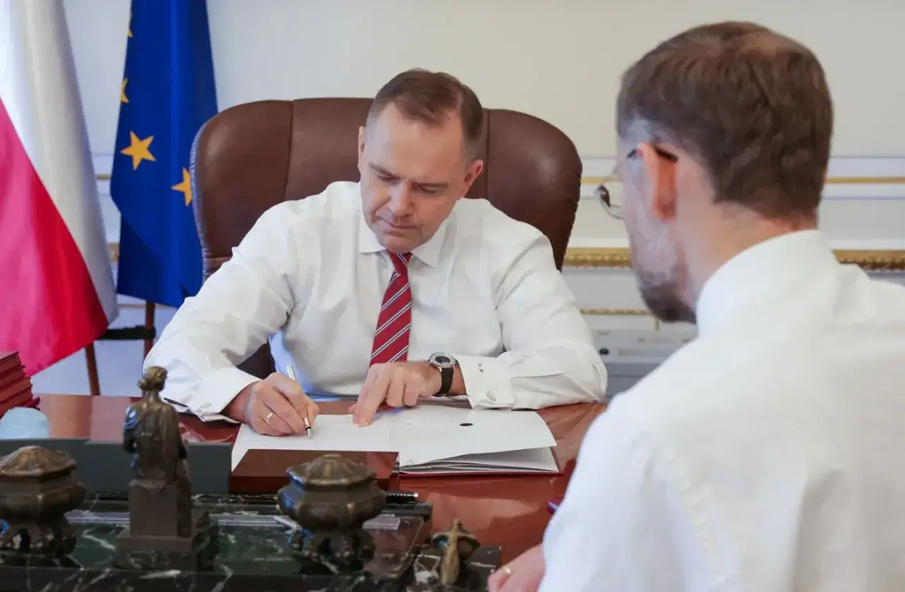 Mężczyzna w białej koszuli i krawacie podpisuje umowę o zlecenie przy biurku, obok flagi Polski i UE.