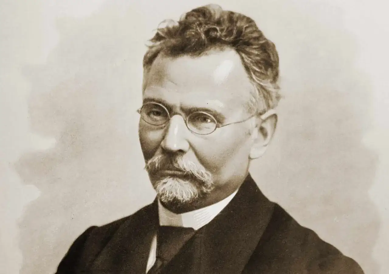Portret mężczyzny w okularach, z wąsami i bródką. Jego twarz przypomina nieco postać z