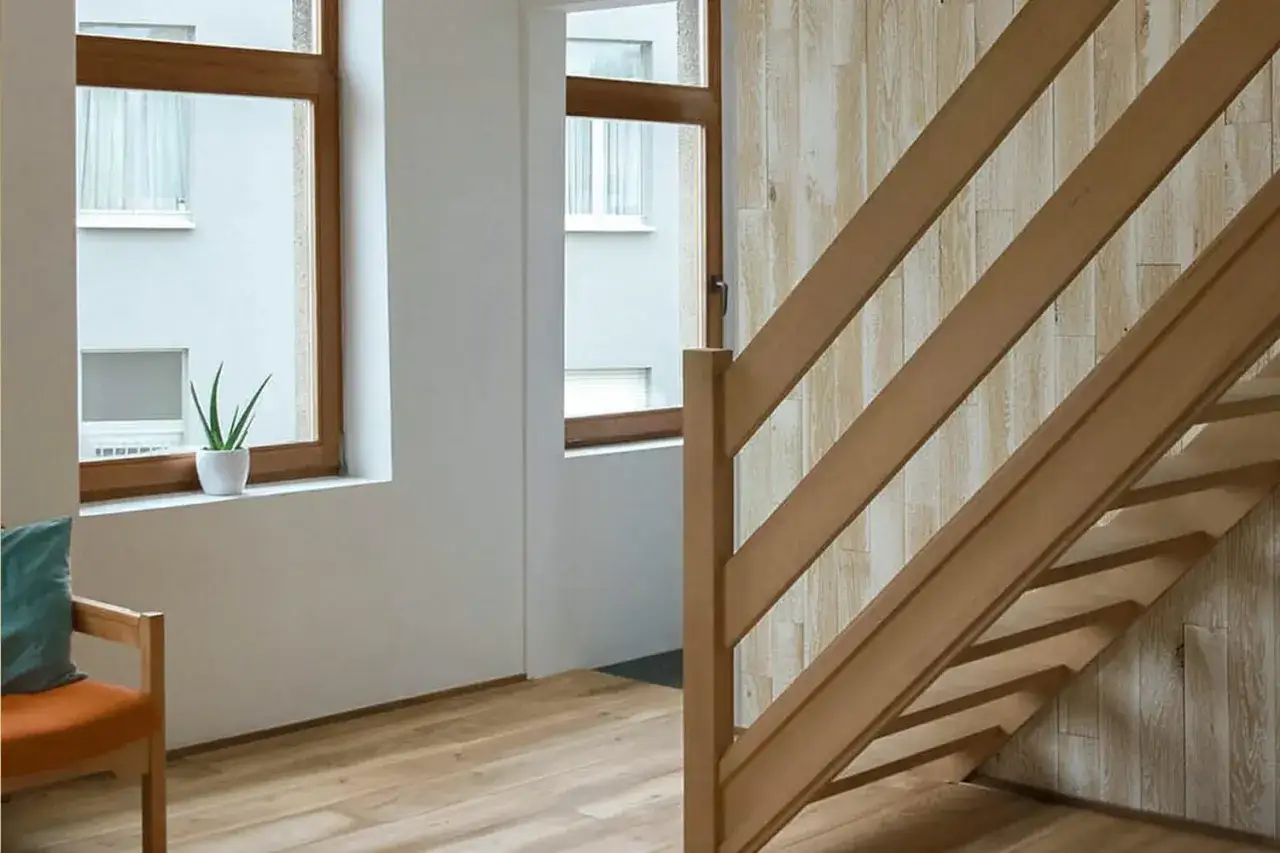Habillage mural textur&eacute; derri&egrave;re une cage d'escalier en bois clair, avec des fen&ecirc;tres et un fauteuil orange.