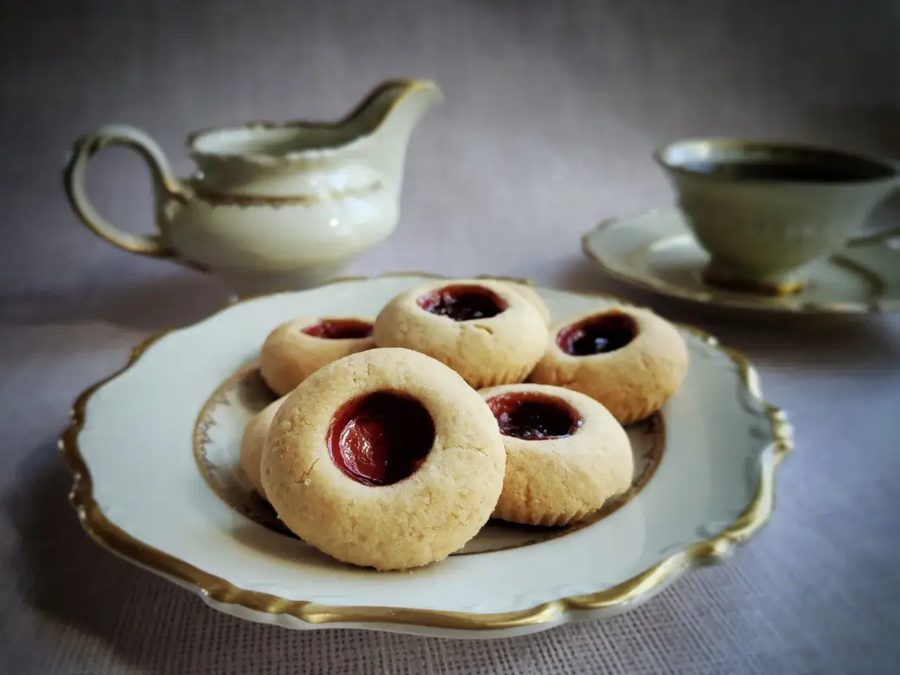 Hallongrottor recept: sm&aring;, runda kakor med hallonsylt i mitten, serverade p&aring; en elegant tallrik med en kaffekopp och gr&auml;ddkanna i bakgrunden.