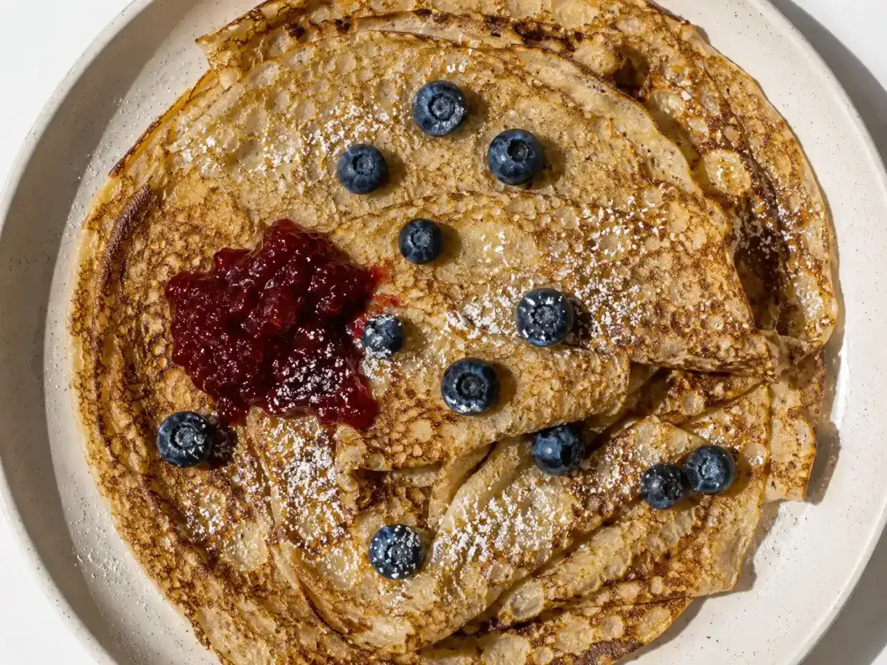 En hög med gyllene, glutenfria pannkakor toppas med färska blåbär, sylt och pudrat socker. En härlig frukost eller fika!