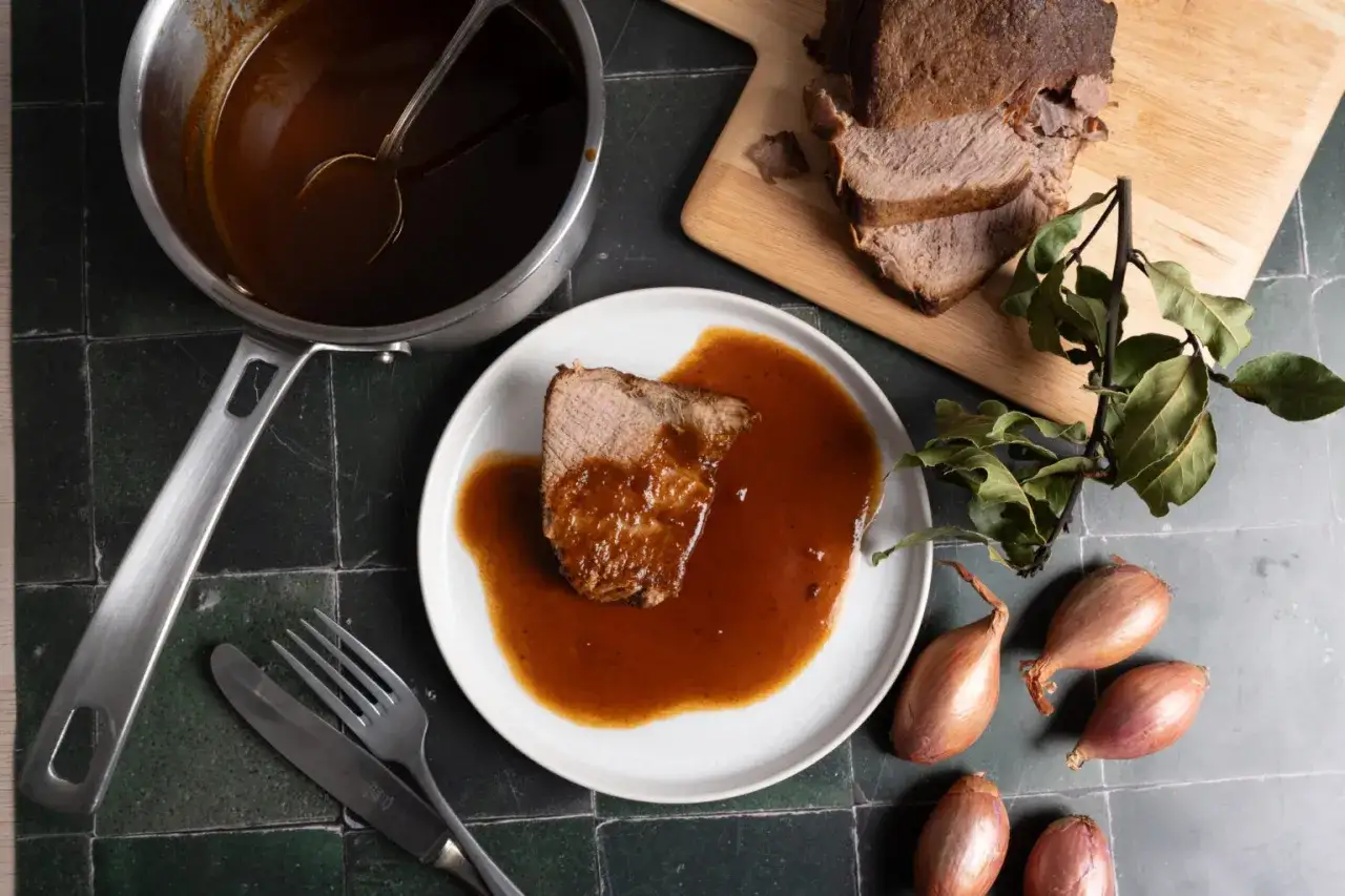 Ein Stück Rinderbraten auf einem Teller, übergossen mit einer köstlichen Rinderbraten Sauce. Daneben ein Topf mit Sauce, Besteck und Schalotten.