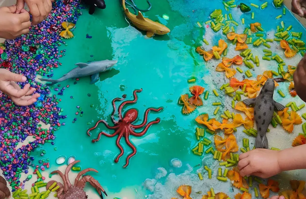 Hands explore a vibrant ocean-themed sensory bin with toy sea creatures, colorful pebbles, and pasta. Perfect for sensory activities for 1 year olds.