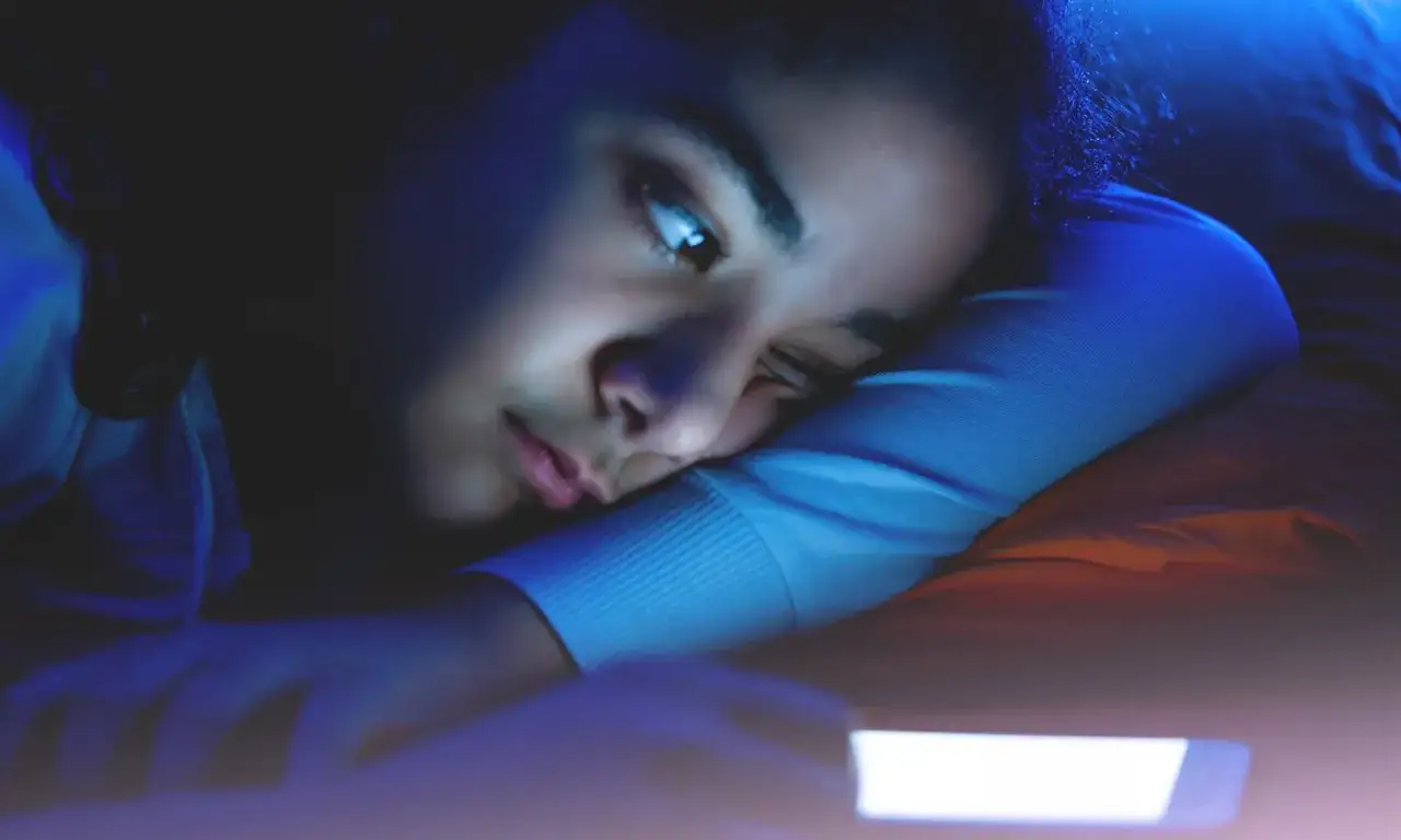 Une jeune fille, éclairée par la lumière bleue d'un téléphone, illustre quelles peuvent être les principales conséquences de la cyberdépendance : isolement, fatigue, et perte de sommeil.