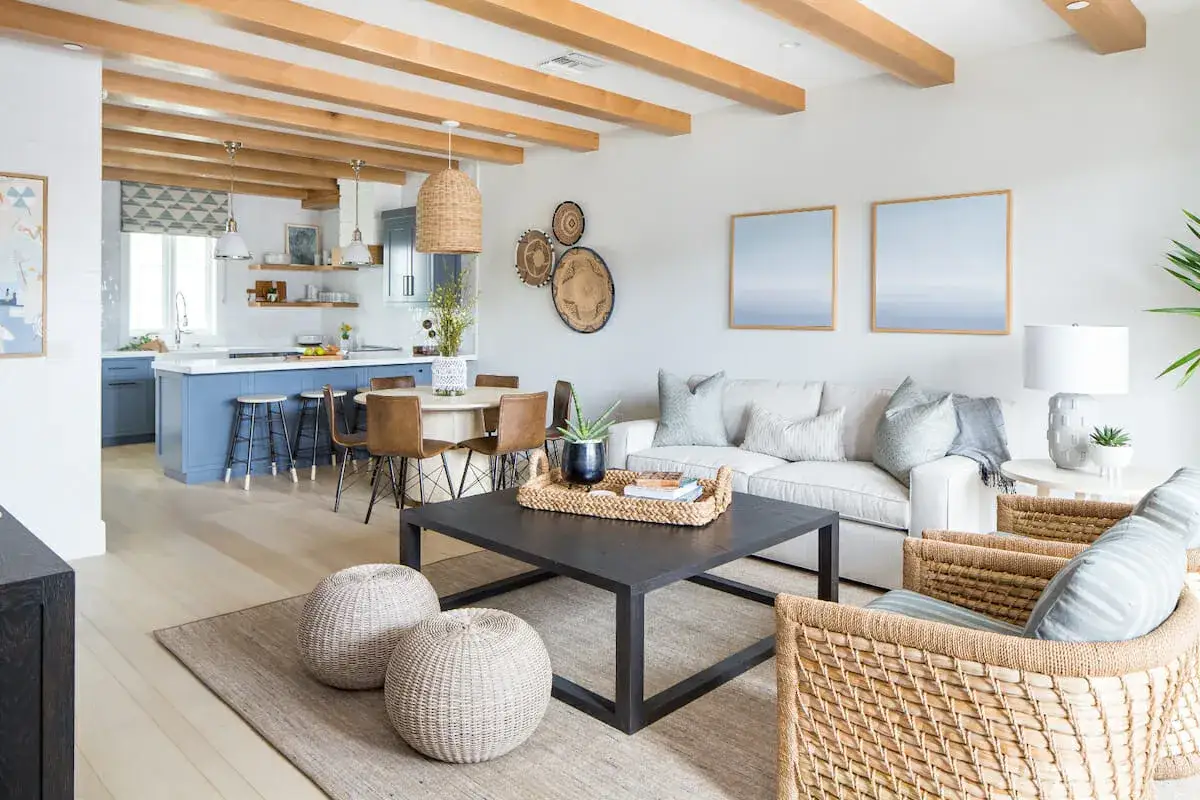 A bright living room with a blue kitchen island, natural wood beams, and woven accents embodies coastal interior design.