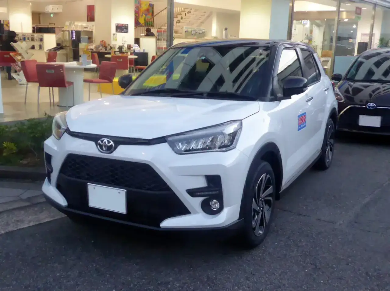 Biały Toyota Raize z czarnym dachem stoi przed salonem samochodowym.