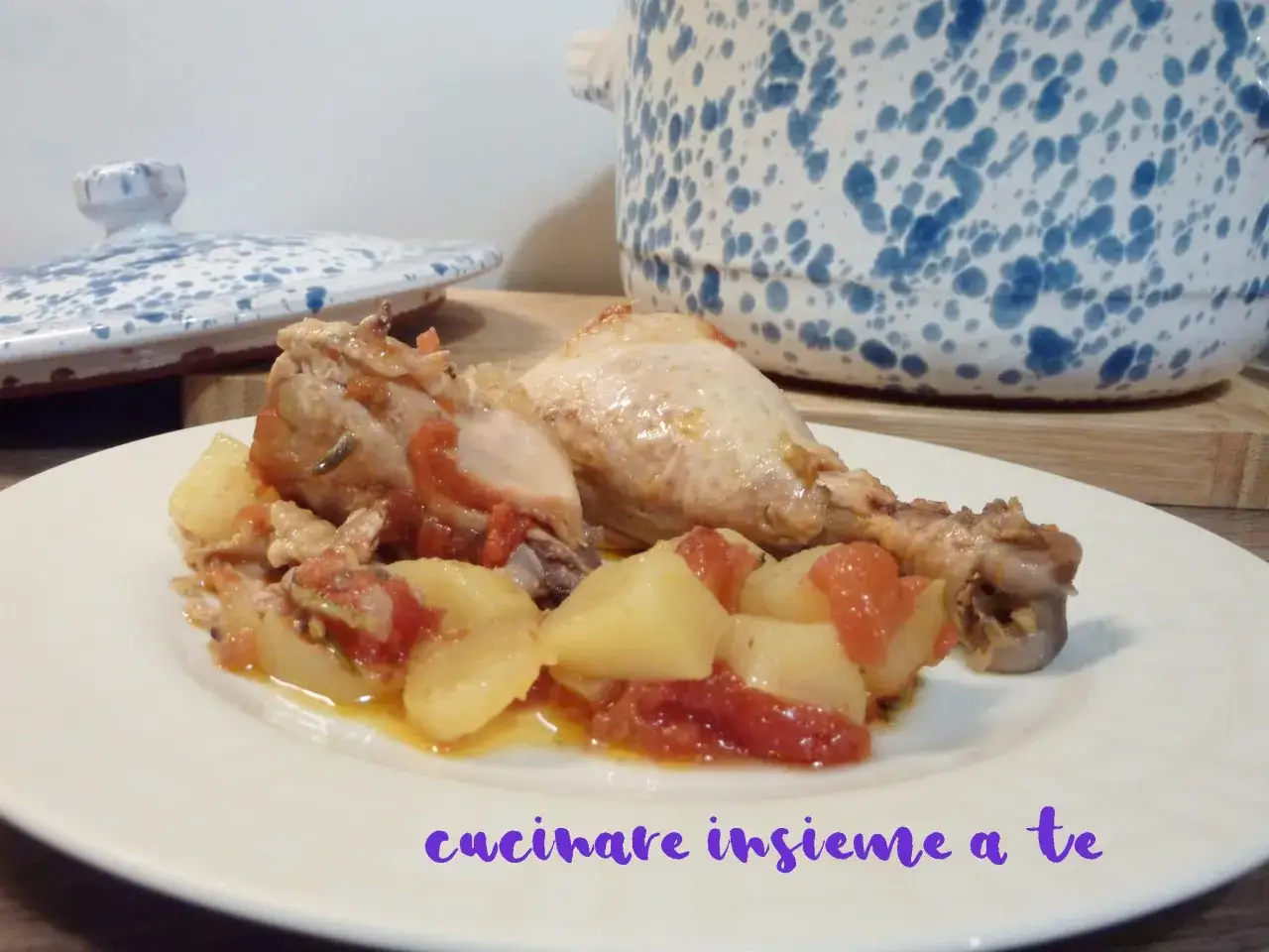 Piatto di pollo in umido con patate e pomodoro, servito in un piatto bianco. Sullo sfondo, una pentola con decorazioni blu.