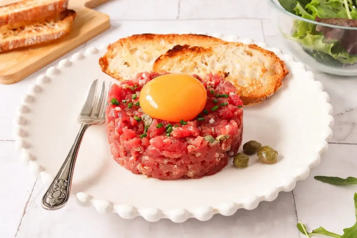 Un delicioso steak tartar con yema de huevo, alcaparras y tostadas crujientes, listo para disfrutar.