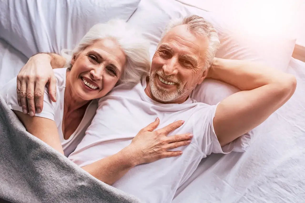 Coppia anziana sorridente a letto, abbracciati. Un esempio di 15 segnali di una relazione sana.