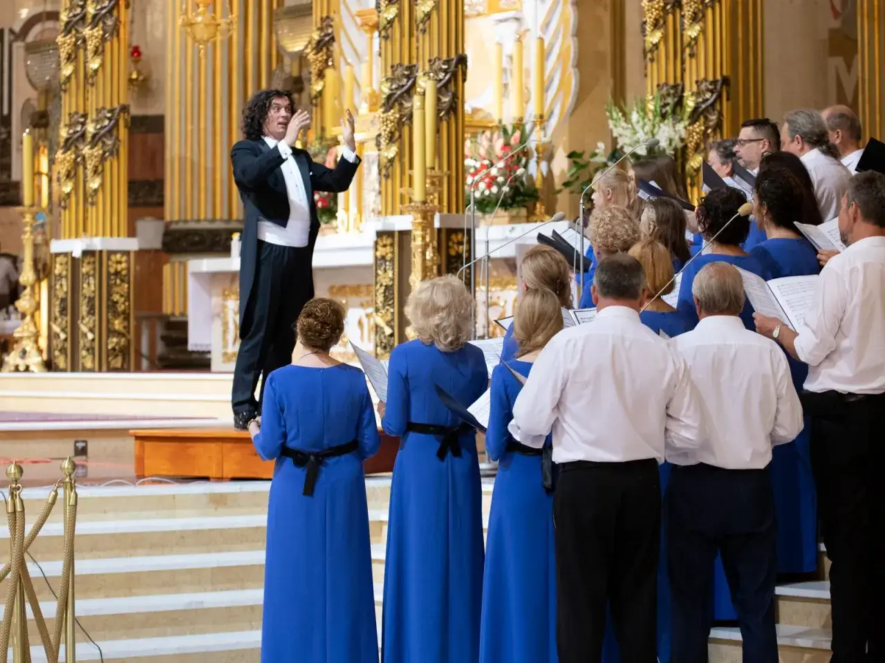 Dyrygent prowadzi chór wykonujący najpiękniejsze pieśni liturgiczne. W błękitnych sukniach i białych koszulach śpiewacy tworzą harmonijną całość.