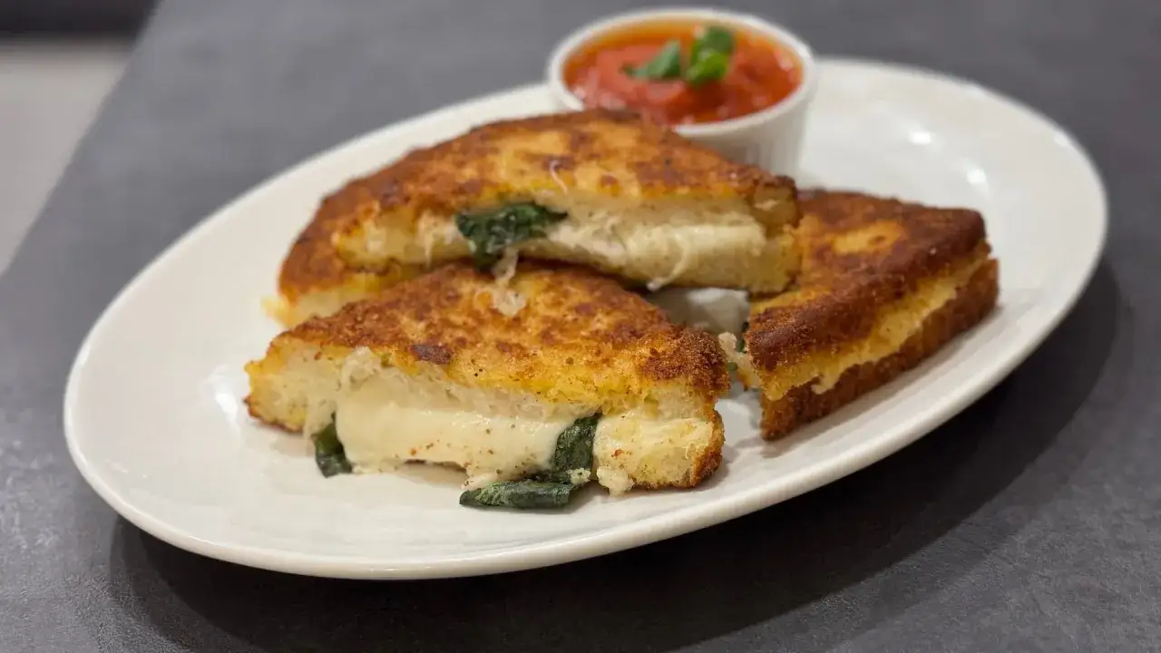 Drei goldbraun gebratene Sandwiches mit geschmolzenem Mozzarella und Spinat, serviert mit einer Schale Tomatensauce.
