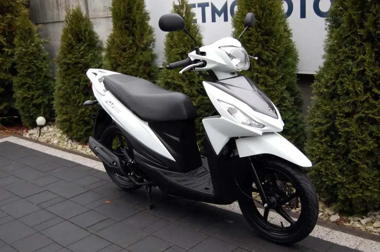 Biały skuter Suzuki Address 110 stoi na ciemnej kostce brukowej, otoczony zielonymi tujami.