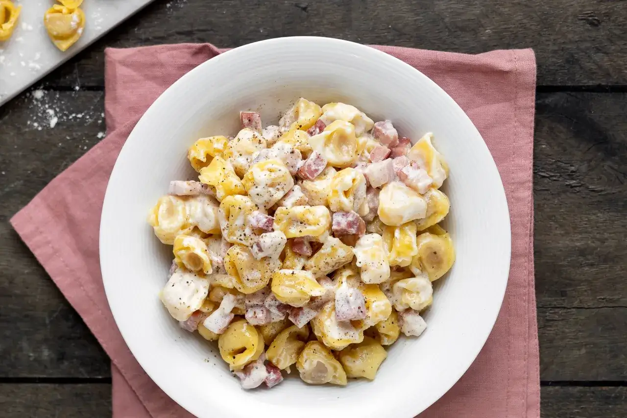 Un piatto di tortellini panna e prosciutto, conditi con una cremosa salsa e pepe nero, serviti su un tovagliolo rosa.