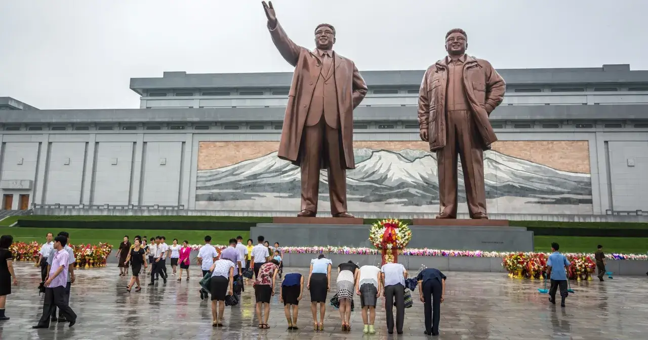 Pomnik przywódców Korei Północnej. W tle budynek, przed nim ludzie kłaniający się. Widać tu wiele zakazów, które obowiązują w Korei Północnej.