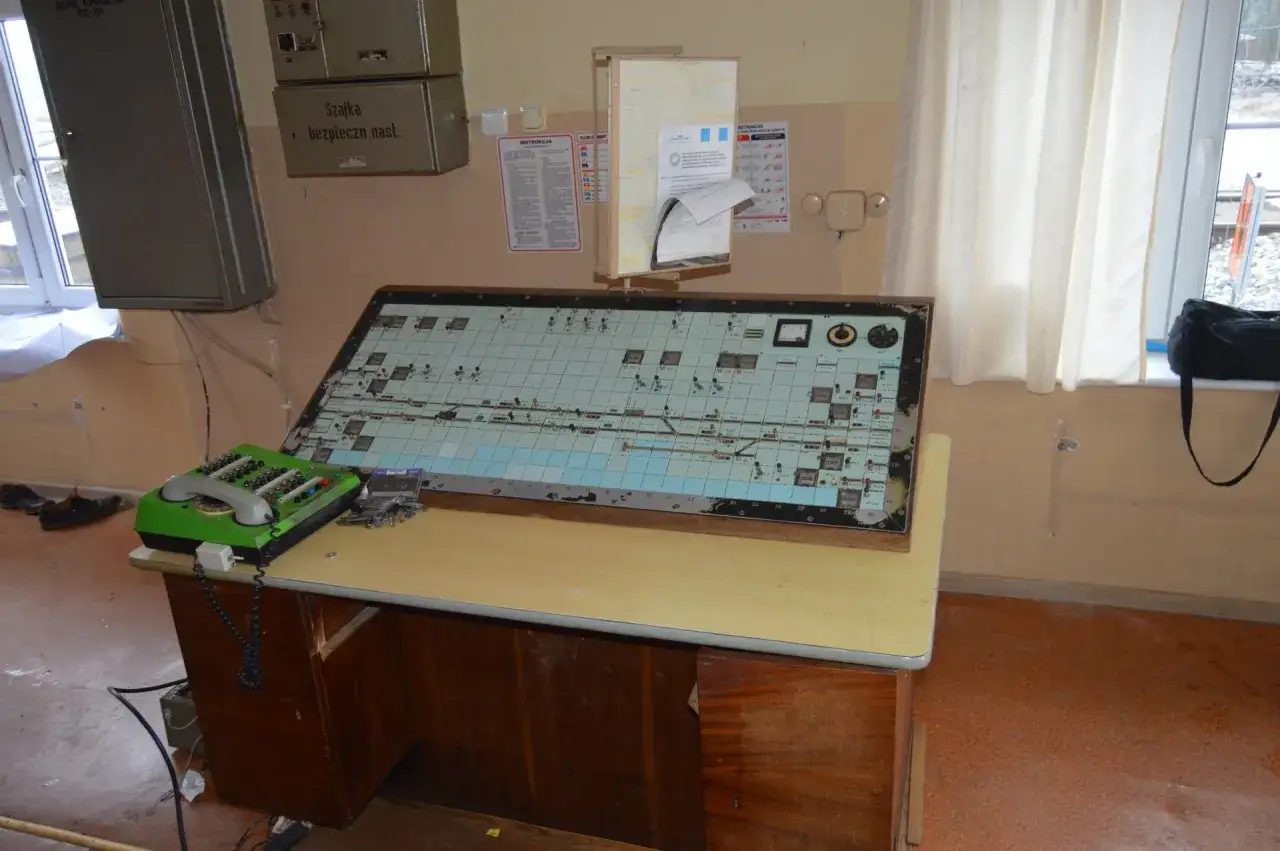 Panel sterowania z mapą tor&oacute;w i przyciskami. Obok zielony telefon i klucze. Na ścianie wisi instrukcja IR-1.