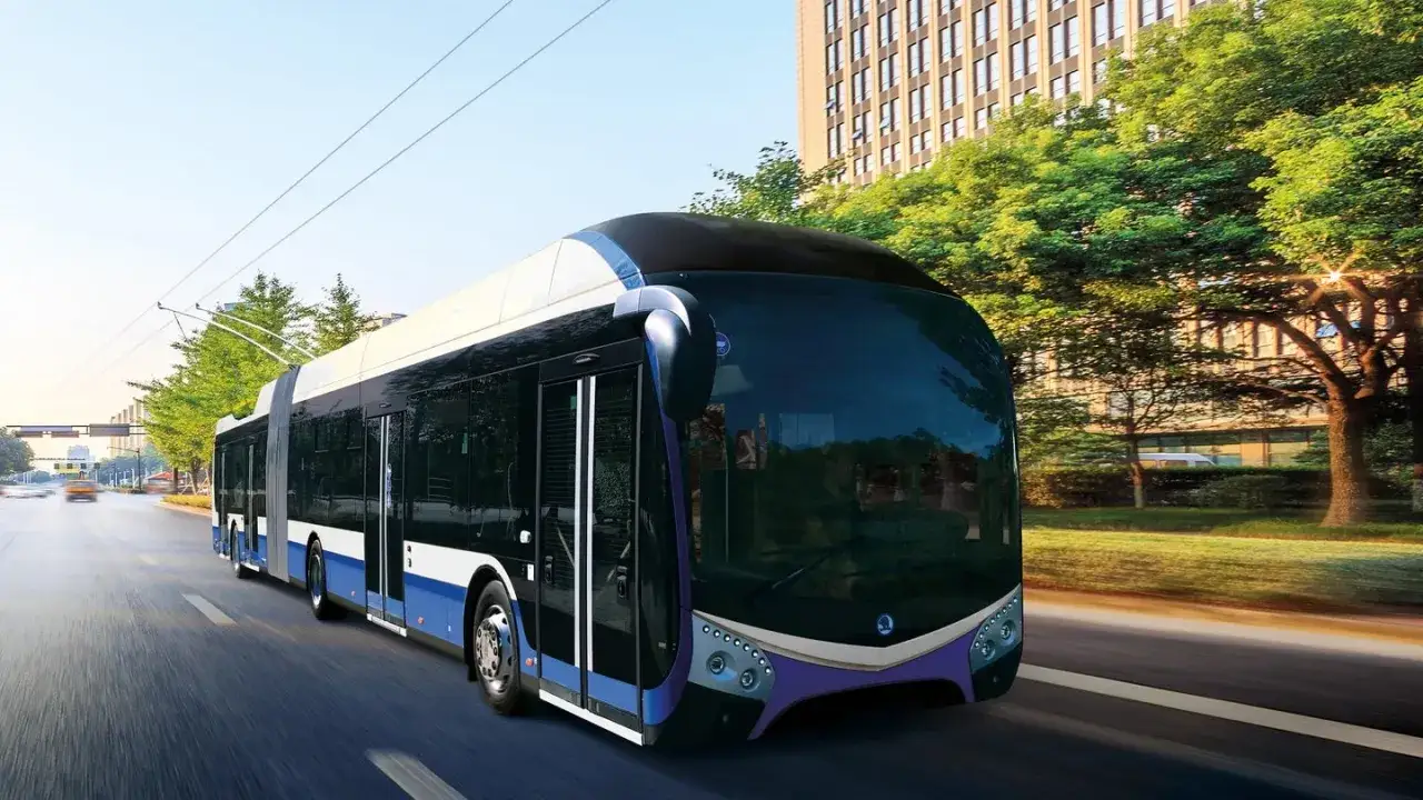 Moderner, langer Gelenkbus f&auml;hrt auf einer Stra&szlig;e an B&auml;umen und Geb&auml;uden vorbei.