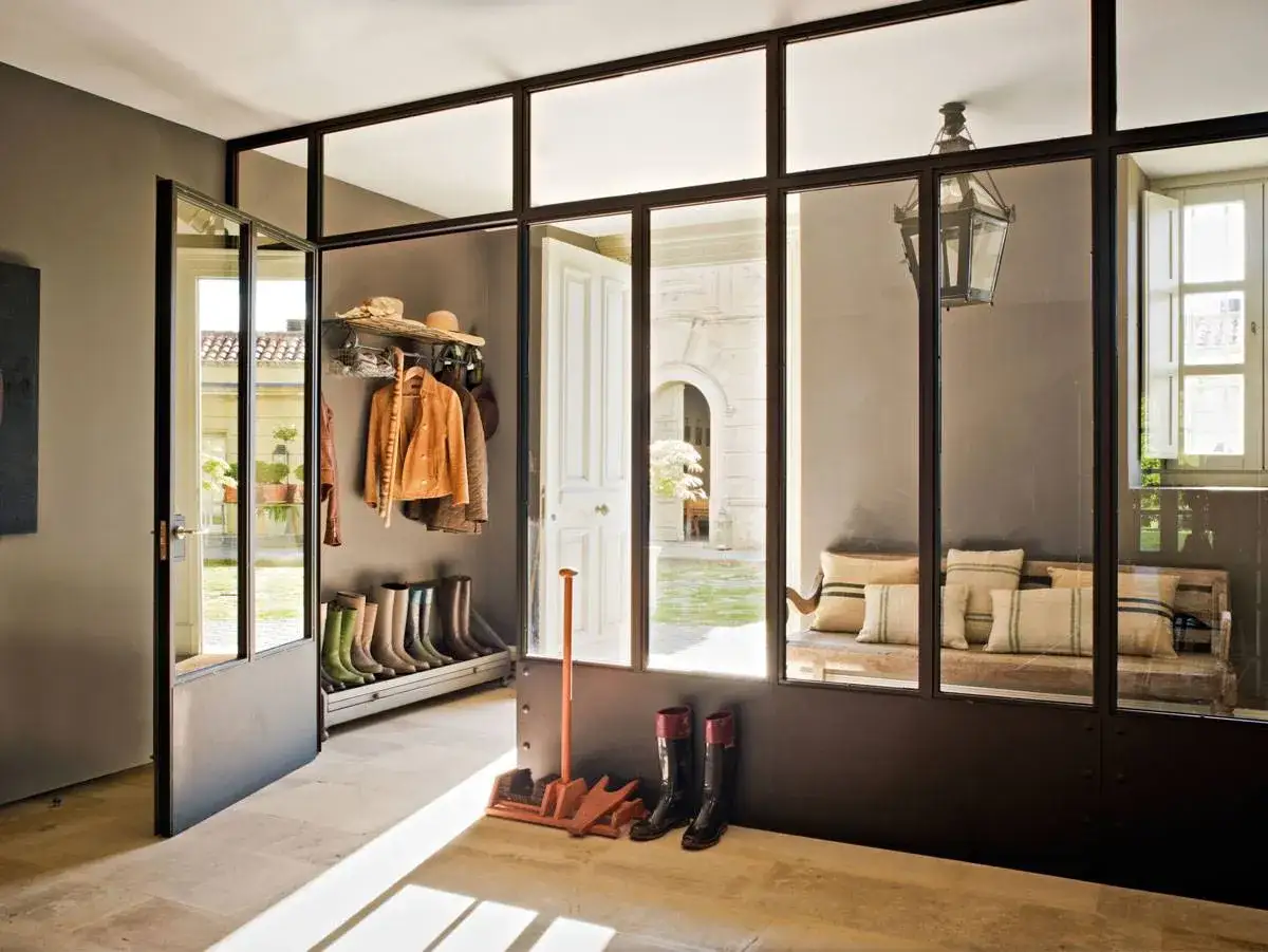 Entrada con botas, chaquetas y sombreros colgados, separada por **paredes de cristal** de un patio interior con banco y cojines.