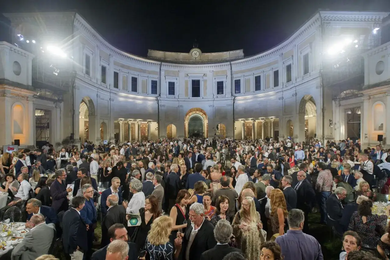 Una folla elegante partecipa a un evento serale all'aperto, con un edificio storico illuminato sullo sfondo. Sembra una celebrazione per il premio Strega.