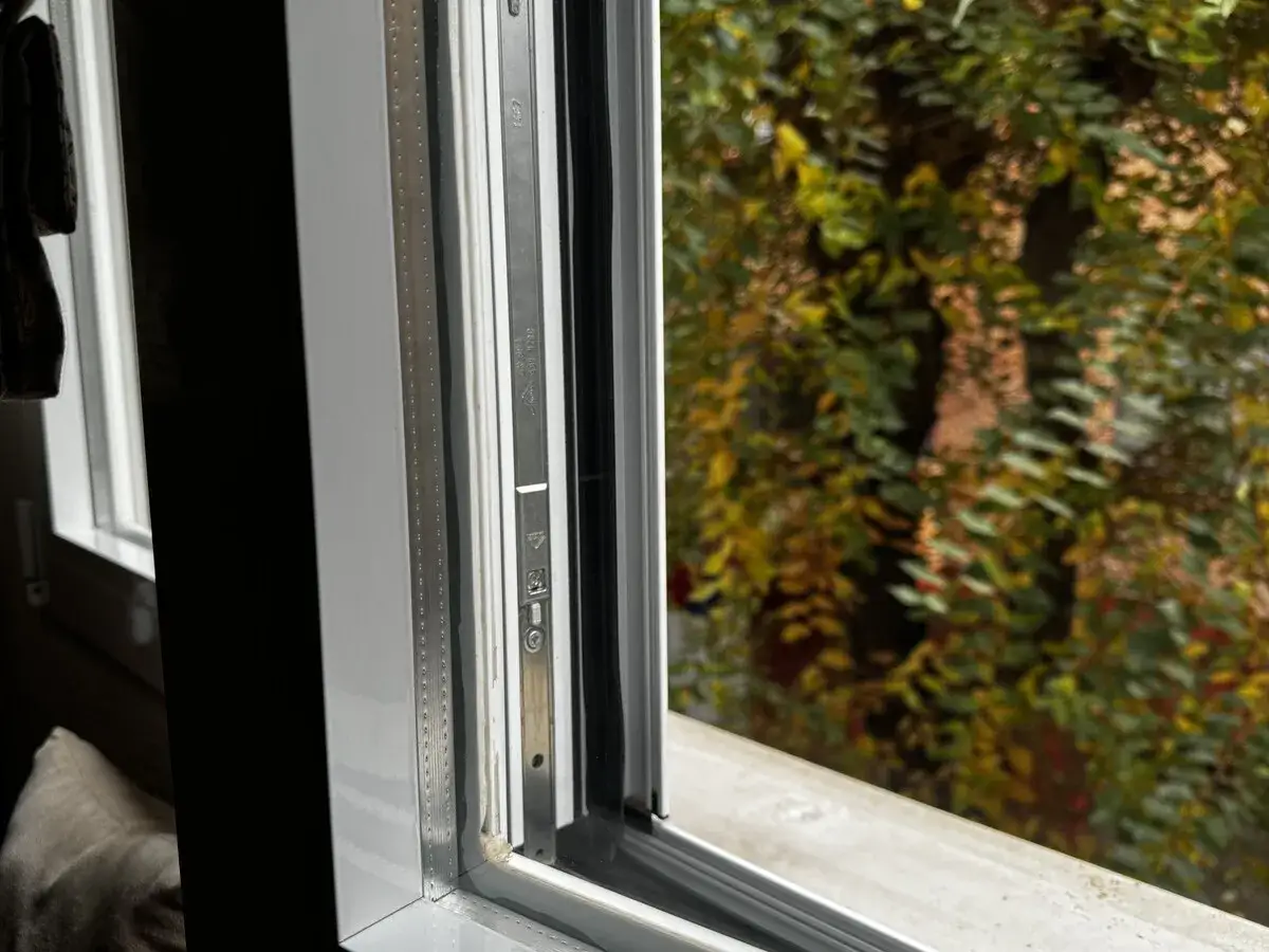 Marco de ventana blanco con detalles met&aacute;licos y vista a un &aacute;rbol frondoso. El **cristal Planitherm** permite disfrutar del exterior.