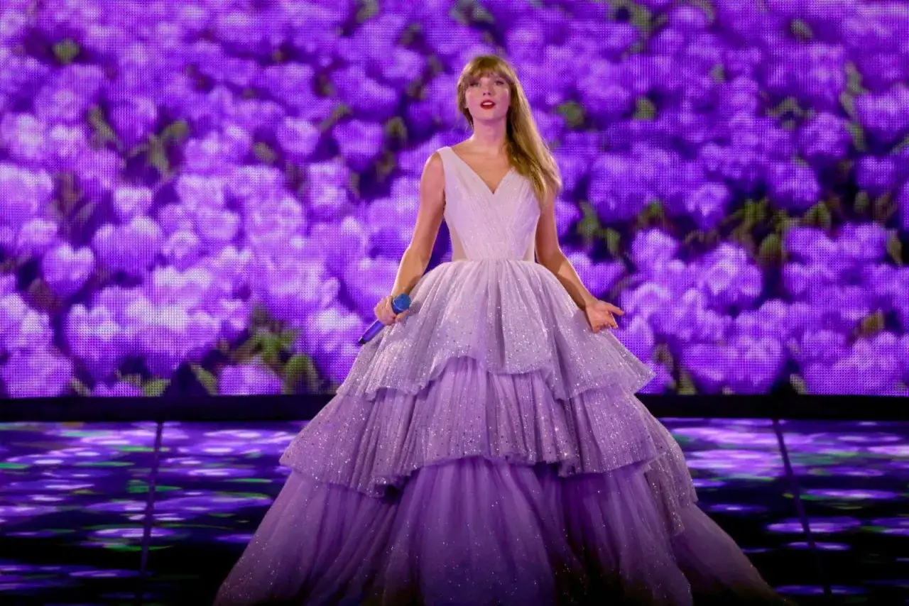 Taylor Swift w fioletowej sukni z falbanami, z mikrofonem w dłoni, śpiewa na tle purpurowych kwiatów. Czas mówić!