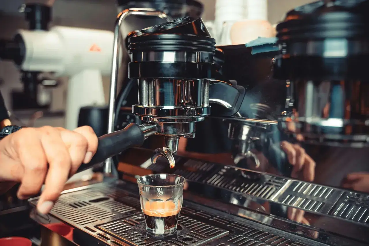 Eine Hand hält den Siebträger einer Espressomaschine, während ein Kaffee lungo in ein Glas läuft.