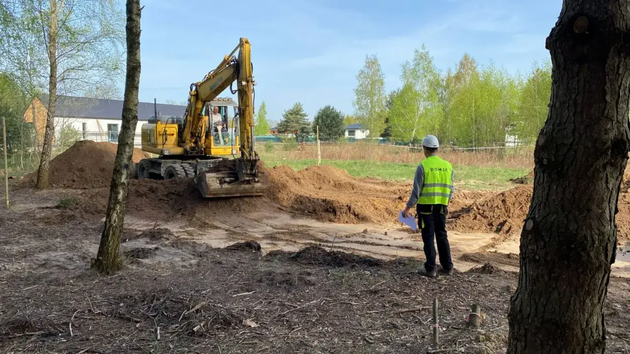 Koparka pracuje na placu budowy, przygotowując działkę pod budowę. Pracownik w kamizelce nadzoruje prace.