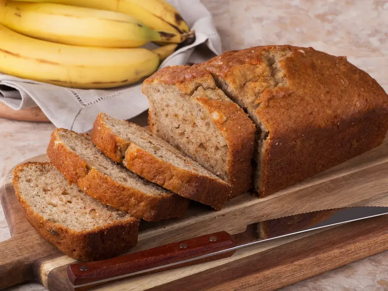Ein saftiges bananenbrot ohne zucker, frisch aufgeschnitten auf einem Holzbrett. Daneben liegen reife Bananen.