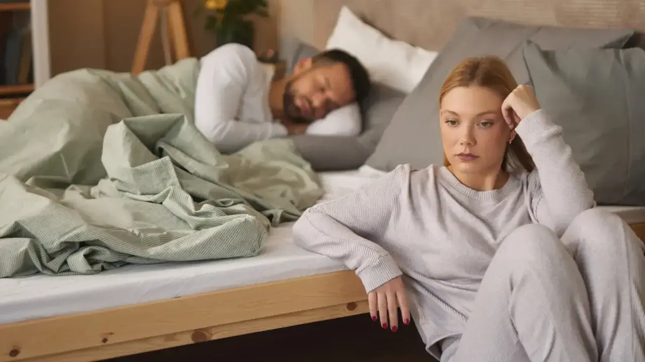 Mujer pensativa sentada en el suelo, mientras su pareja duerme en la cama. Reflexiona sobre qué significa soñar que tu pareja te es infiel.