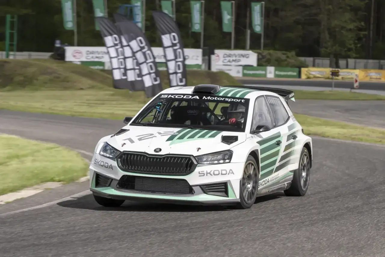 Biała Skoda Fabia Rally z zielonymi pasami pędzi po torze. Kierowca w kasku jest gotowy do akcji.