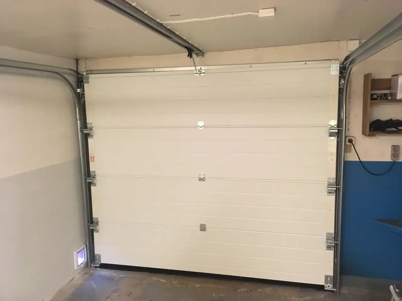 Monterad verisure magnetkontakt på garageporten. En vit sektionsport med metallskenor och gångjärn.