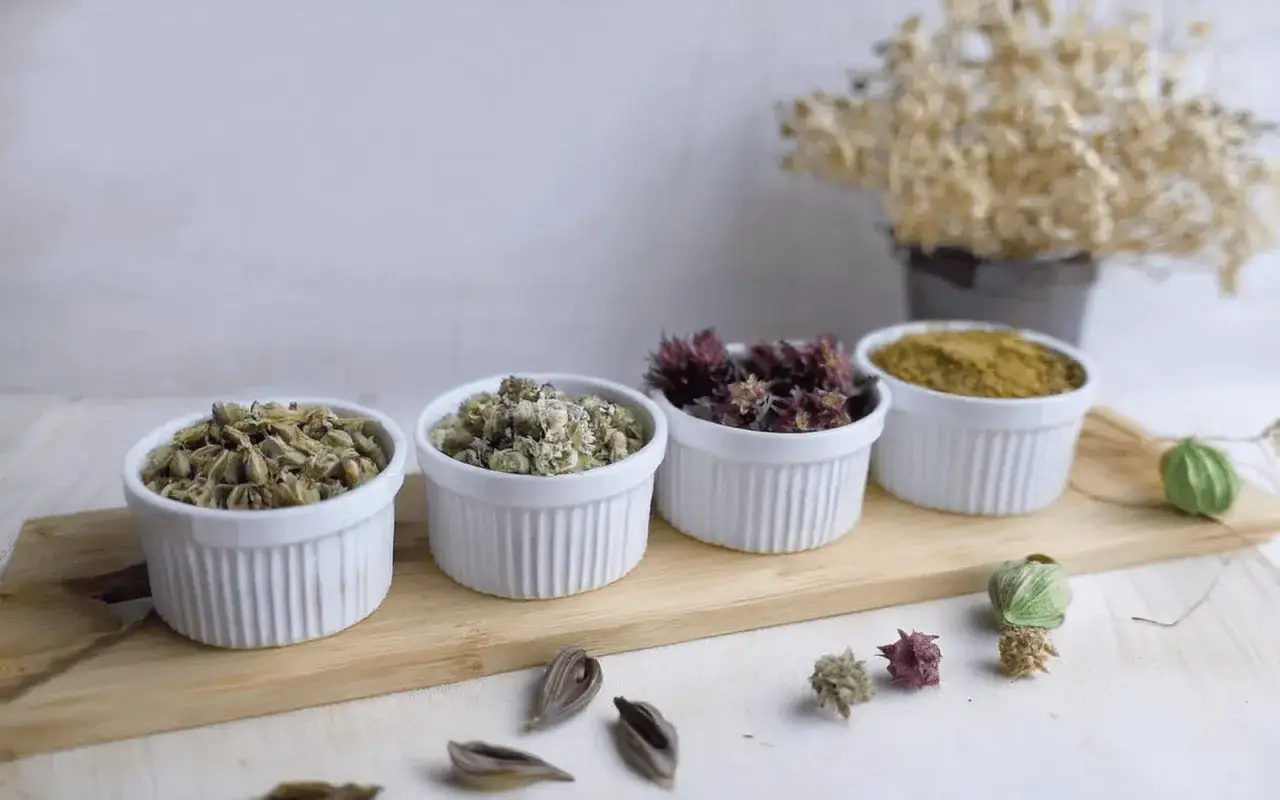 Quattro ciotole bianche con erbe essiccate, ideali per una tisana per digerire. Sul tavolo, fiori secchi e semi.