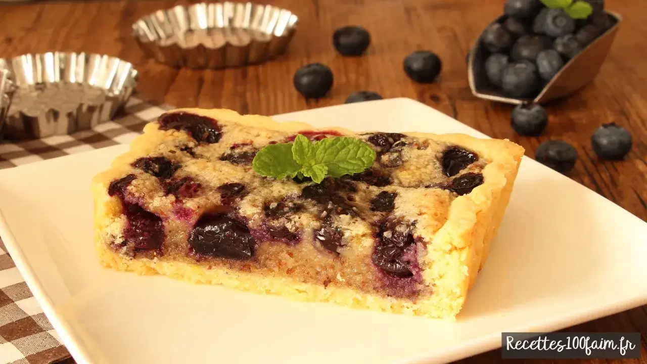 Tranche de tarte myrtille amande, garnie d'une feuille de menthe, sur une assiette blanche. Des myrtilles fraîches parsèment la table en bois.