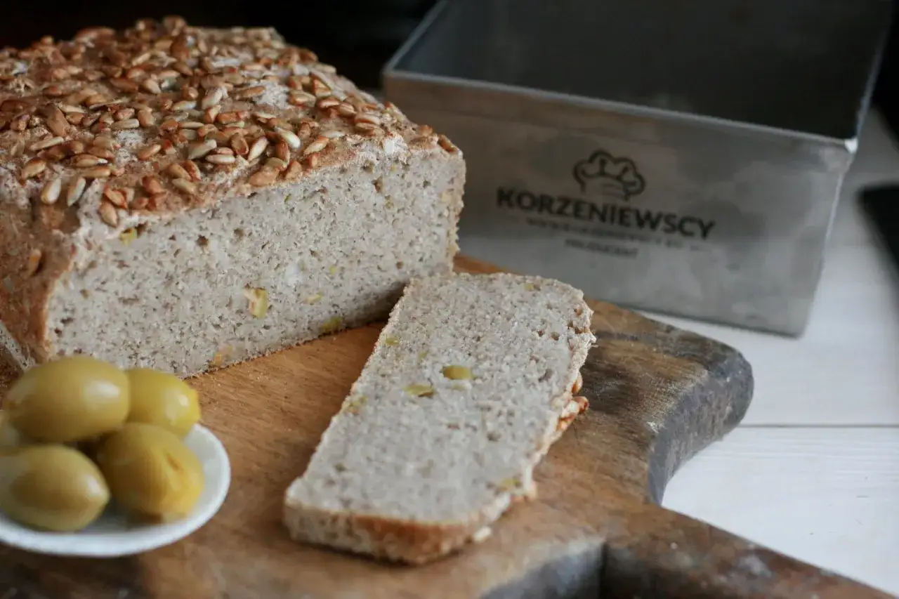 Pyszny chleb bezglutenowy posypany słonecznikiem, obok kromka i oliwki. Obok forma do pieczenia.