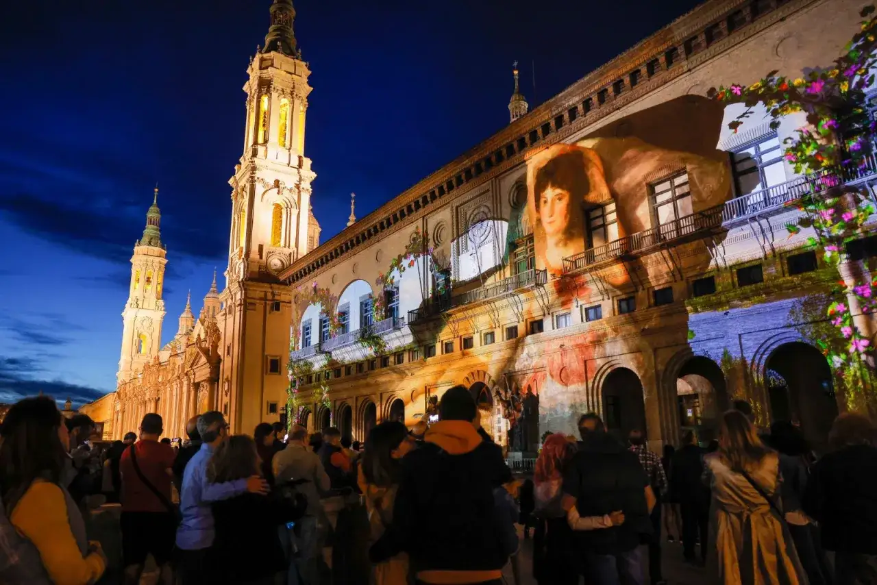 Proyección de arte en la fachada de un edificio histórico durante las fiestas goyescas, con gente observando.