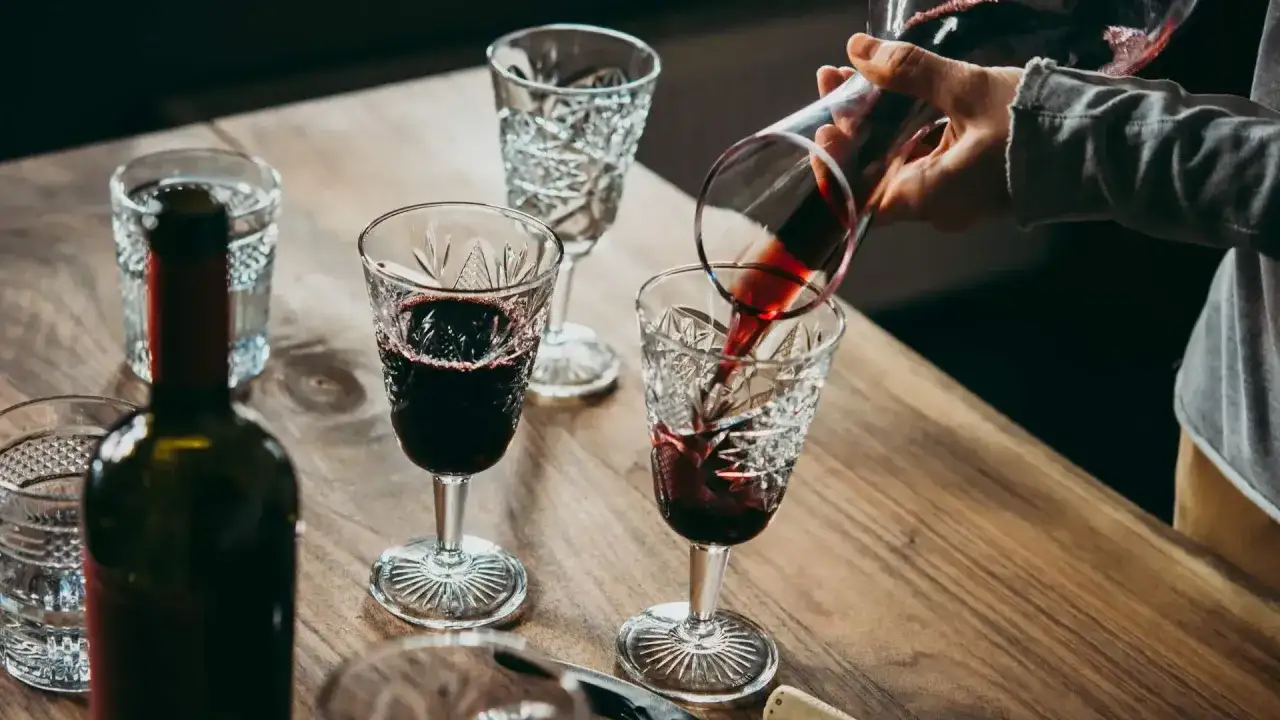 Czerwone wino nalewane do kieliszka, obok butelka i inne kieliszki. Osad w winie widoczny na dnie.